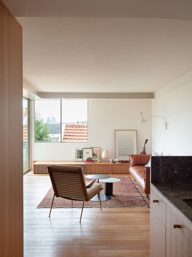 A warm, refined open-plan living area with light timber flooring and built-in timber joinery. The room features a cognac leather sofa, mid-century lounge chair, nesting coffee tables, a patterned area rug, artwork, and a sculptural floor lamp. Large windows provide abundant daylight and views toward neighbouring rooftops and a distant city skyline. A dark stone kitchen benchtop and timber cabinetry are visible in the foreground.
