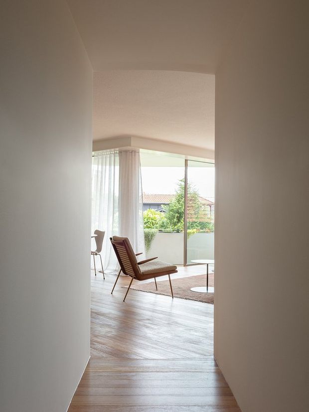View through a plain hallway into a bright mid-century-inspired living room. The space features light timber flooring, floor-to-ceiling glazing with sheer curtains, a brown upholstered lounge chair, a small round table, and a balcony or terrace beyond. Neutral painted walls and a softly textured ceiling create a calm, airy atmosphere.