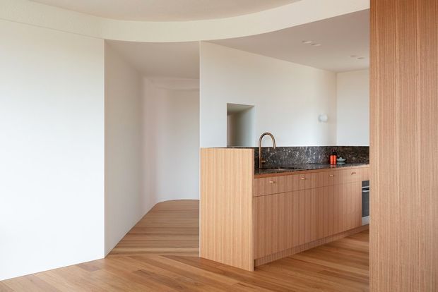 Minimalist open-plan kitchen with warm vertical timber cabinetry, a dark stone benchtop and integrated appliances. The kitchen is set against smooth white walls with a curved ceiling bulkhead and adjacent timber flooring extending into a hallway or living area.
