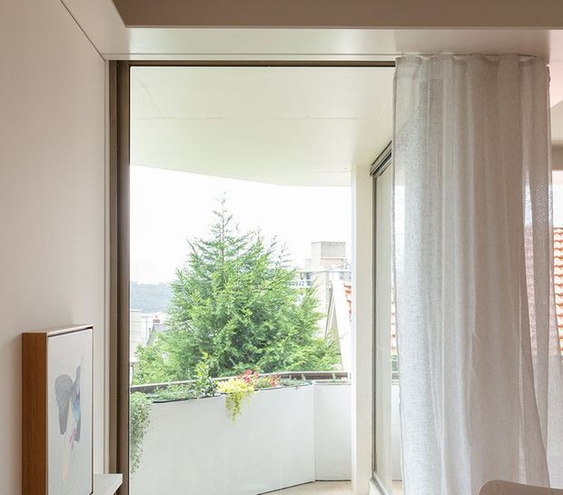 Bright interior view through a large glazed opening to a small planted balcony or terrace. A sheer white curtain hangs at right, with pale walls, a timber-toned window or door frame, and a leafy residential outlook beyond.