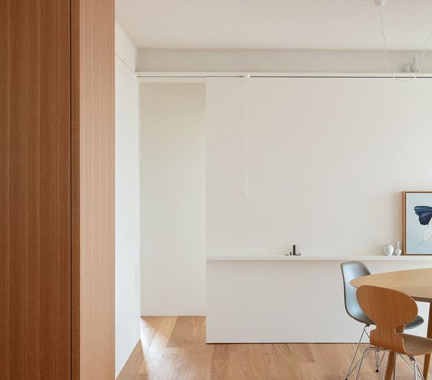 Minimalist open-plan kitchen and dining area with warm timber flooring and vertical timber wall panelling. A sleek white kitchen island with integrated cabinetry and a small black tap sits against white walls, with sculptural modern dining chairs and pendant lighting visible at the right.
