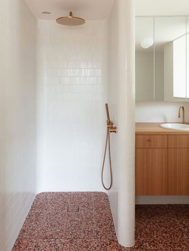 A refined contemporary bathroom featuring an open walk-in shower lined with slim white vertical tiles, a terrazzo shower floor, and warm brushed brass shower fittings. A curved tiled partition partially separates the shower from a timber vanity with an integrated white basin, mirrored cabinetry, and matching terrazzo flooring.