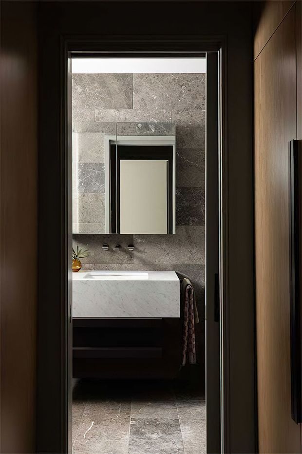 A dark, refined bathroom viewed through an open doorway. The room features large grey veined stone wall and floor tiles, a white marble-look vanity basin with dark timber cabinetry, black-framed mirror and fixtures, and warm timber wall panelling at the doorway. A small plant and hand towel add minor accents.
