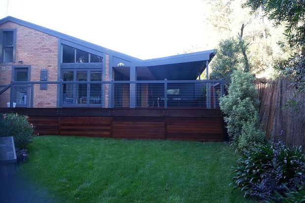 Exterior view of a contemporary single-storey home with a strong gabled brick section, extensive dark-framed glazing, and a covered outdoor terrace. The building sits above a timber-clad deck or platform with horizontal metal balustrading, opening onto a small landscaped lawn and enclosed garden.