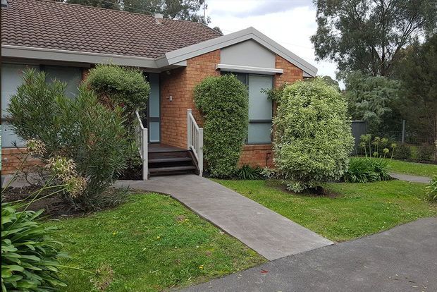 Single-storey suburban brick dwelling with a tiled pitched roof, grey gable trim, dark-framed windows, a small covered entry porch and concrete path. Mature shrubs and lawn surround the frontage, with a paved driveway or walkway visible in the foreground.