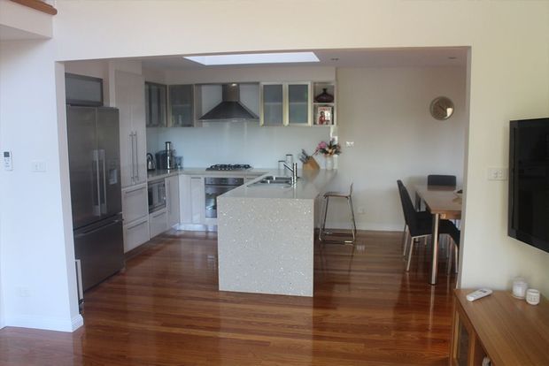 Open-plan kitchen and dining area with polished timber flooring, white cabinetry, stainless-steel appliances, a central stone island, and a skylight. A dining table sits beside the kitchen, with a television and low timber console visible at the right.