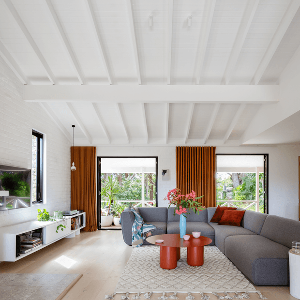 Bright contemporary living room with a large grey sectional sofa, red cylindrical coffee tables, patterned rug, white-painted brick walls, and a striking white exposed-beam ceiling. Full-height windows and glazed doors open to leafy greenery, while rich vertical timber curtains add warmth and texture.