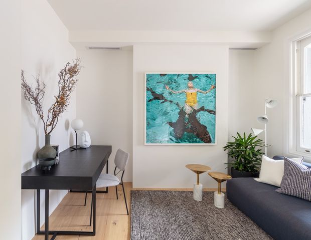 Bright contemporary living room with white walls, light timber flooring, a dark minimalist console desk and chair, charcoal sofa, textured grey rug, indoor plant, sculptural side tables, and colourful artwork. Large windows provide abundant natural light.