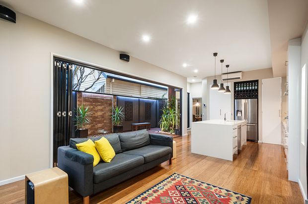 Open-plan contemporary living and kitchen area with a dark charcoal sofa, patterned rug, white kitchen island, pendant lights, timber flooring, and extensive glazed sliding doors opening toward a planted courtyard. Warm timber and neutral finishes are accented by bright yellow cushions and a colourful geometric rug.