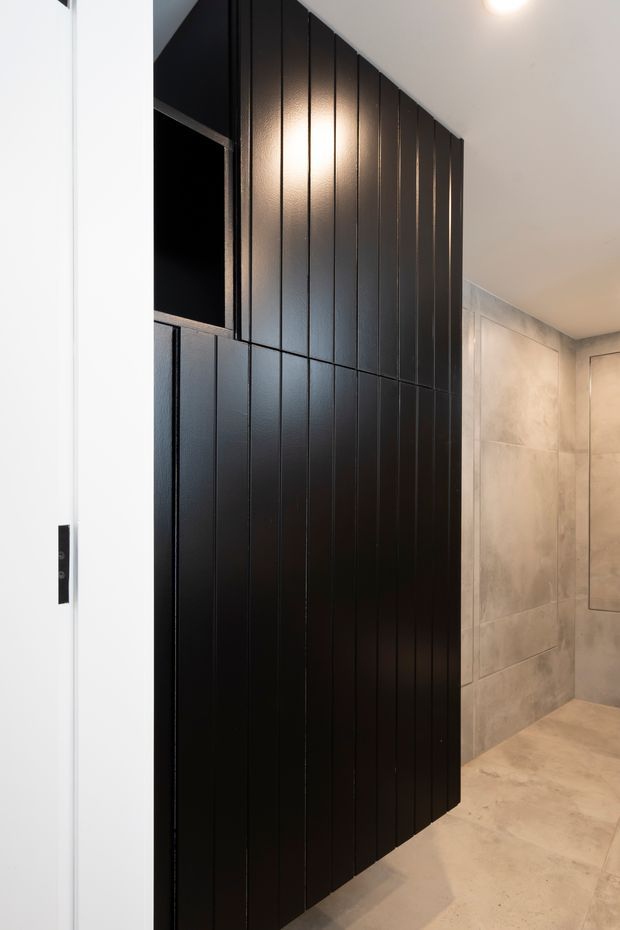 A contemporary interior passage or bathroom area featuring a dramatic black vertically fluted wall or fixed screen with an open recessed niche. Pale large-format stone-look wall and floor tiles form the surrounding space, with warm recessed lighting and a clean minimalist finish.