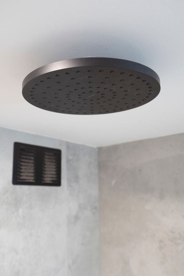 Close-up view of a round matte black rainfall showerhead mounted to a white ceiling in a minimalist bathroom shower enclosure. The surrounding walls have large grey concrete-look finishes, with a black rectangular ventilation grille visible on the wall.