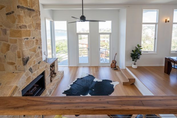 Bright open-plan living room with a substantial rustic stone fireplace, warm timber flooring, white walls, and tall windows and glazed doors overlooking a coastal or urban landscape. A contemporary ceiling fan, minimalist furnishings, guitar, indoor plant, and dark hide rug add relaxed character. A timber stair or balustrade crosses the foreground.