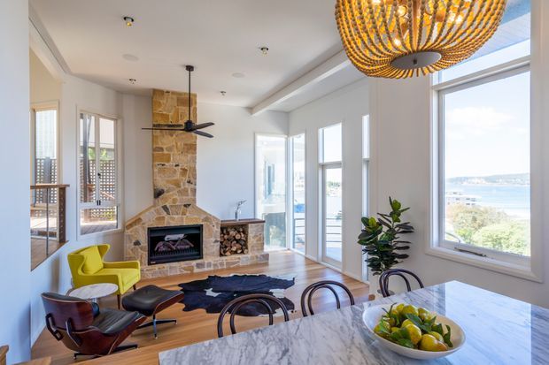 Bright, airy open-plan living and dining area with a prominent natural stone fireplace, timber flooring, large coastal-facing windows, sculptural pendant lighting, and a marble-topped dining island or table. Mid-century-style lounge chairs and contemporary furnishings create a refined coastal atmosphere.