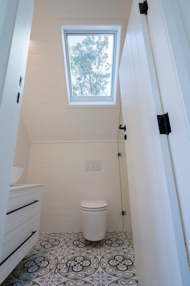 Bright compact bathroom viewed through the open doorway, featuring a white toilet, white vanity with black pulls, white horizontal wall panelling, patterned decorative floor tiles, and a skylight-style window bringing in natural light. The door and hardware are white with black hinges and fittings.