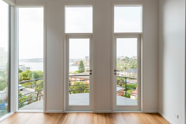 Bright, minimal interior room with pale walls, warm timber flooring, and three tall glazed openings, including two narrow glazed doors, overlooking a leafy residential neighbourhood and water beyond. Dark metal balcony railings are visible outside. The room is unfurnished and appears to be a living or general residential space.