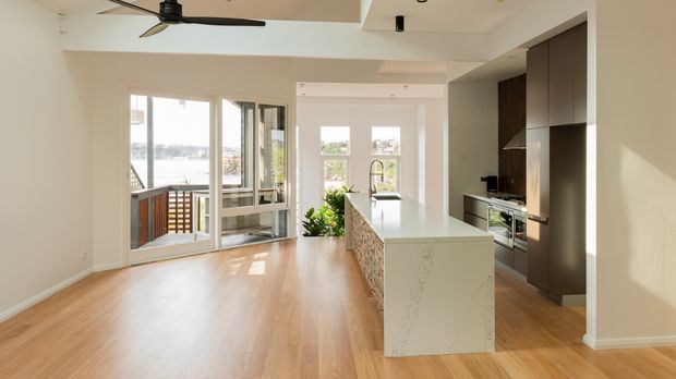 Bright open-plan kitchen and living area with light timber flooring, a large pale stone island, dark full-height cabinetry, stainless-steel appliances, and a ceiling fan. Glazed doors open to a balcony with a distant water view, while strong daylight fills the space.