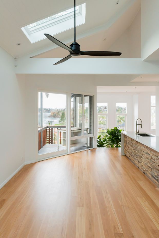 Bright open-plan interior with pale timber flooring, white walls, a high ceiling with skylight and a dark ceiling fan. A compact kitchen island with a stone-faced base and white benchtop occupies the right side, while large glazed doors open to a balcony with a visible water and urban outlook. Lush indoor planting adds a natural accent.