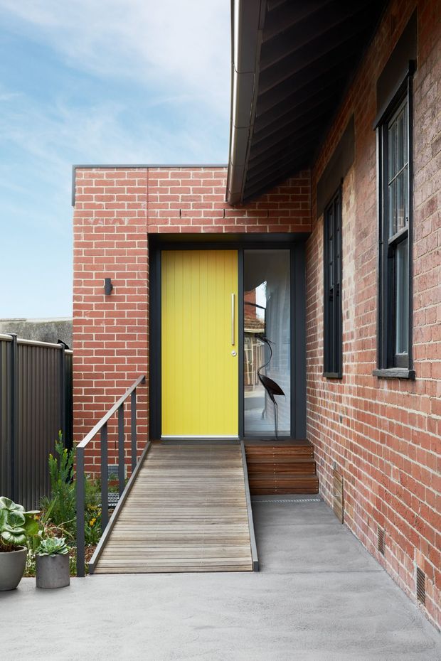Exterior entry approach to a contemporary brick home, featuring a bright yellow timber front door with sidelights, dark metal window and door frames, a timber ramp and steps, concrete paving, and a simple metal balustrade. Potted plants sit beside the entry, while the projecting roof eave creates a sheltered walkway.
