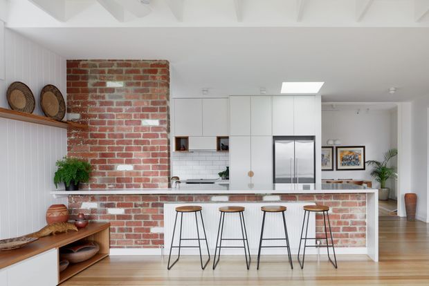 Bright open-plan kitchen with a large central island clad in exposed red brick and topped with a pale benchtop. White flat-panel cabinetry, white subway-tile splashback, stainless-steel appliances, timber flooring, and a white panelled ceiling create a clean contemporary setting. Four black-framed timber-seat stools line the island, with open shelving, plants, artwork, and a small adjoining living or circulation area visible beyond.