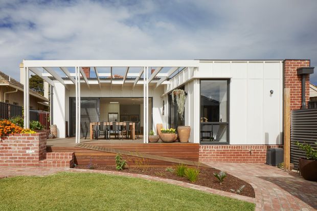Rear exterior view of a contemporary single-storey home extension with a white-framed glazed living or dining area opening onto a timber deck. A pergola-like roof structure provides partial shade, while red brick walls, paved paths, raised garden beds, and landscaped lawn frame the façade.