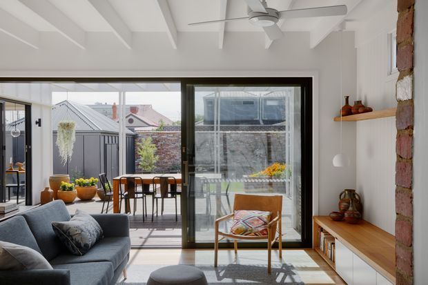 Bright contemporary living room with a large black-framed glazed opening to a small outdoor courtyard or terrace. The room features a grey sofa, timber lounge chair, low timber-and-white built-in media cabinetry, floating timber shelf, exposed brick side wall, white painted walls, and a white beamed ceiling with ceiling fan. Dining furniture and outdoor planting are visible beyond the glazing.