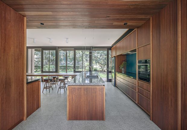 Contemporary kitchen viewed through a timber-lined opening, featuring extensive dark timber cabinetry, a central island with a polished stone benchtop, integrated appliances, a deep blue-green splashback, and full-height glazing overlooking trees. The ceiling and surrounding walls are also clad in warm timber, while the floor is a neutral grey finish. A dining table and chairs sit beyond the island.