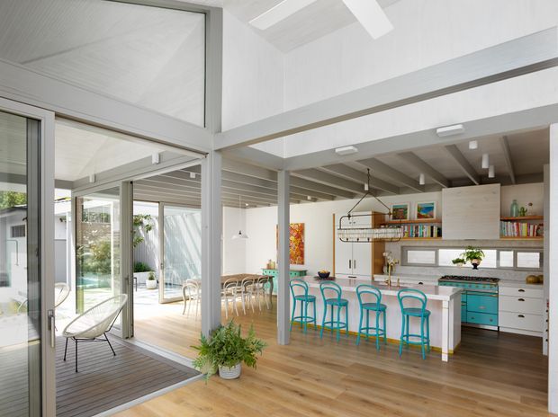 Bright open-plan residential interior with a kitchen and casual dining area opening onto a covered outdoor deck through extensive sliding glass doors. The space features light timber flooring, white painted walls and exposed structural beams, a large island with a pale stone-look benchtop, turquoise bar stools, white cabinetry, open shelving, and abundant natural light. A high ceiling and clerestory-like glazing create an airy, connected indoor-outdoor feel.