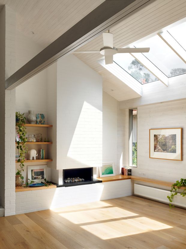 Bright contemporary living room with white-painted brick walls, a white timber-lined ceiling, exposed dark structural beam, skylights, and a large white ceiling fan. Built-in open timber shelving, a linear fireplace, window seat, radiator, framed artwork, and pale timber flooring create an airy, warm interior.