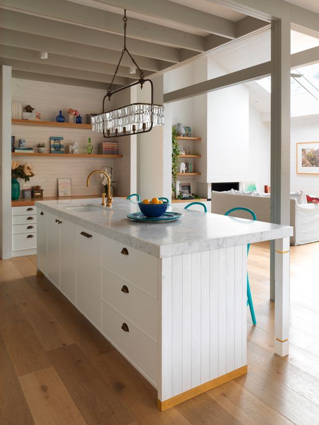 Bright, open-plan kitchen centred on a large white island with a pale stone benchtop, shaker-style drawers, cup pulls, and vertical white panelled sides. Warm timber flooring, open timber shelves with decorative objects, a brass gooseneck tap, and a statement hanging chandelier define the space. Large white-framed glazing and a partial living area are visible beyond the kitchen.