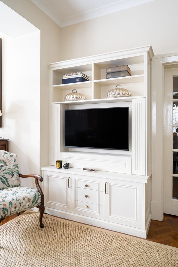 Bright traditional living room featuring a custom white built-in entertainment unit with paneled cabinetry, open display shelving, and an integrated television. The room has warm cream walls, white decorative trim, a patterned upholstered armchair, and a natural woven rug over dark timber flooring. A glazed door is visible at the right.