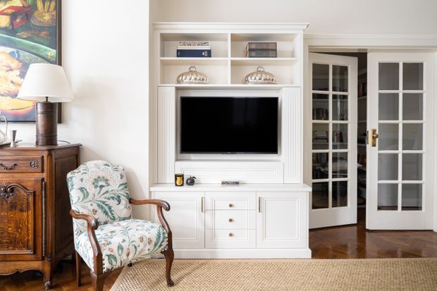 A refined traditional living room featuring a white built-in entertainment centre with open shelving, framed cabinetry, and a recessed television. The room includes a carved dark-wood sideboard, a floral upholstered armchair, a table lamp, framed artwork, white French doors with divided glazing, warm parquet flooring, and a natural woven rug.