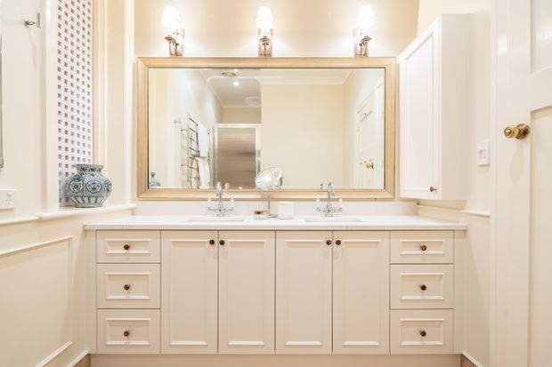 Bright traditional bathroom vanity with a long cream-painted double-sink cabinet, white benchtop, framed wall-to-wall mirror, and polished chrome fittings. Symmetrical wall sconces illuminate the space, while raised-panel cabinetry, brass door hardware, and classic trim create a refined, period-inspired look.