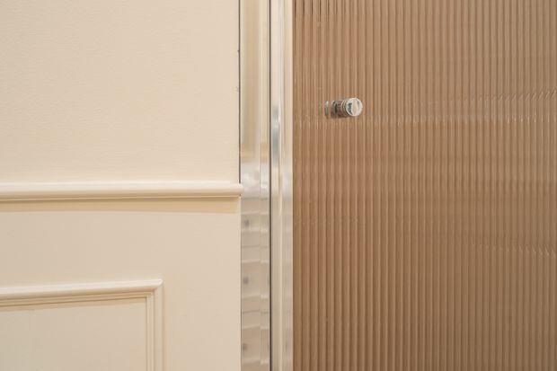 Close-up detail of a refined interior doorway or shower enclosure, showing cream painted wall panelling with raised moulded trim beside a vertically ribbed warm taupe-brown glass or panel surface. A narrow reflective metallic frame and small round hardware provide a subtle contemporary accent.
