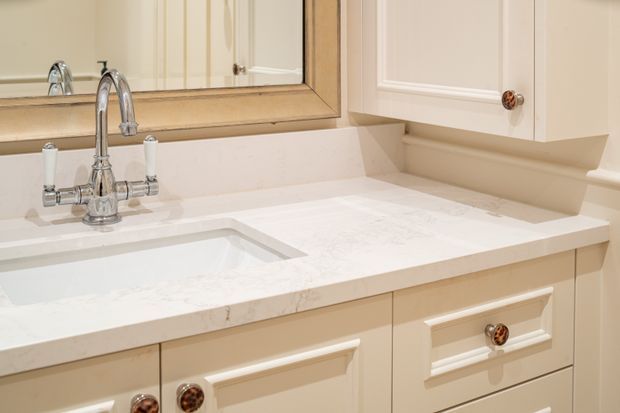 Close-up of a traditional bathroom vanity with a white integrated rectangular basin, polished chrome bridge-style tapware, white veined stone benchtop, cream raised-panel cabinetry, decorative brass-gold knobs, and a framed mirror. The styling is warm, refined, and classic.