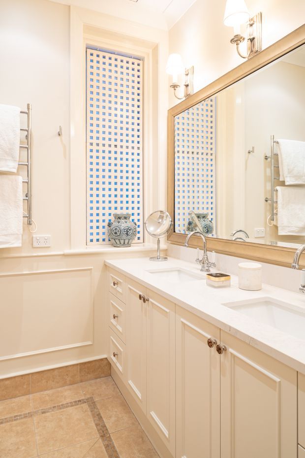 Bright, traditionally styled bathroom vanity with a long cream-painted cabinet, white countertop, dual sinks, polished chrome faucets, framed wall mirror, and twin wall sconces. A tall window with a blue-and-white square-patterned privacy screen provides a decorative focal point. Beige tiled flooring and cream wall panel mouldings complete the warm, refined interior.