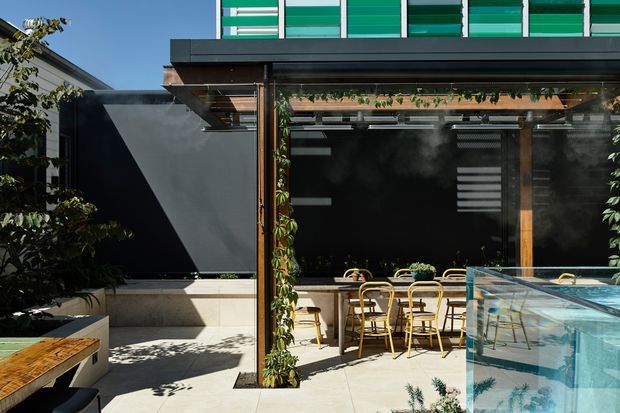 A contemporary outdoor entertaining area with a covered pergola, timber posts, climbing greenery, dark feature walls, and a dining setting. A transparent glass-sided pool or water feature occupies the foreground, while a modern building facade with green horizontal screening is visible above.