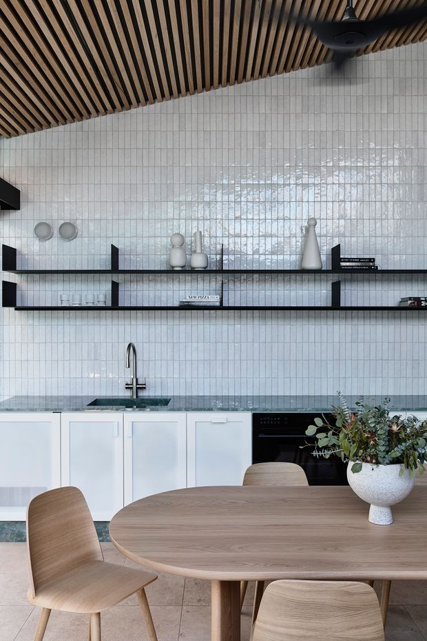 A contemporary kitchen and dining area with white square tiled walls, white lower cabinetry, dark stone benchtops, open black shelving, and integrated black appliances. A warm timber slatted ceiling with a ceiling fan contrasts with the cool tiled surfaces. A rounded timber dining table and matching chairs occupy the foreground, with simple decorative objects and foliage adding understated detail.