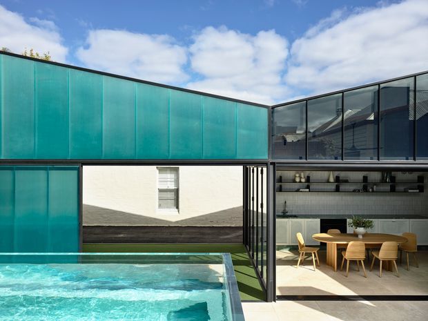 Contemporary courtyard-facing home with a turquoise translucent-panel facade, expansive black-framed glazing, and an open-plan dining area with built-in shelving. A swimming pool occupies the foreground, while a white rendered wall and planted roof edge frame the bright outdoor setting.