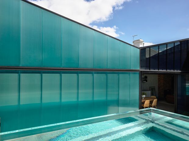 A contemporary courtyard-style building with translucent turquoise panel cladding, dark-framed glazing, and a rooftop swimming pool in the foreground. An open terrace or living area is visible behind the pool, with exposed brick and a chimney adding contrast to the modern envelope. Bright daylight and blue sky create a crisp, airy atmosphere.