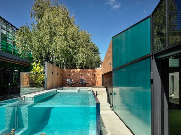 A contemporary courtyard pool enclosed by dark-framed glazing, translucent turquoise screening, timber fencing and warm brick walls. The clear turquoise pool dominates the foreground, with a narrow concrete-paved walkway along the right and mature greenery beyond the courtyard.
