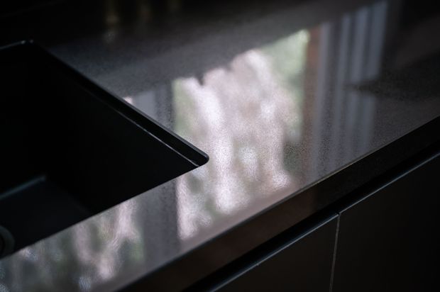 Close-up of a dark, glossy kitchen benchtop with a black inset sink or cooktop visible at the left and dark cabinetry below. Reflections from a window create soft highlights across the polished surface.