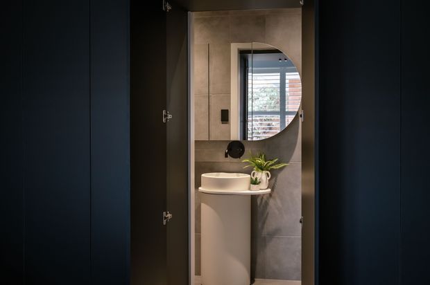 A compact bathroom viewed through partially open dark cabinetry or a concealed doorway. The bathroom features large grey concrete-look wall tiles, a rounded white vessel basin on a slim pedestal vanity, a black wall-mounted tap, a circular mirror, and a small potted plant. Natural light enters through a window with horizontal blinds, creating a moody, refined contemporary setting.