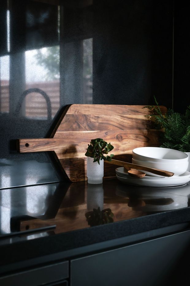 A moody close-up of a contemporary kitchen or dining preparation area, featuring a polished dark stone benchtop, dark cabinetry, a large natural timber serving board, stacked white ceramic bowls and plates, wooden utensils, and greenery. Soft daylight enters from the left and is reflected across the glossy surfaces.