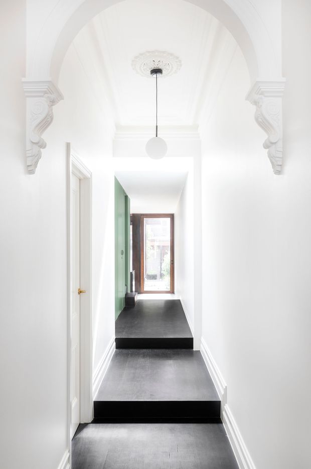 A bright, narrow residential hallway with stepped dark flooring, white walls and ornate classical plaster detailing. A large arched opening with decorative corbels and ceiling medallion frames the corridor, which leads toward a timber-framed glazed door. A green built-in or wall surface appears along the left side, and a simple spherical pendant hangs centrally.