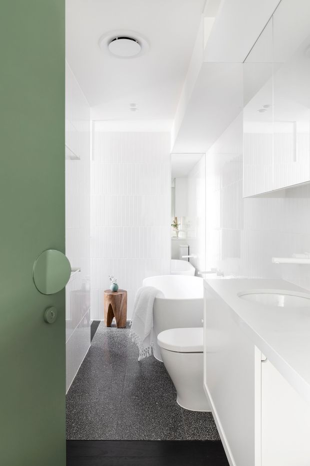 Bright, narrow contemporary bathroom viewed through a muted green door. The room features white vertically textured wall tiles, a white freestanding bathtub, toilet, integrated vanity with basin and mirrored cabinetry, dark terrazzo-like flooring, and minimalist recessed lighting. A small timber stool and simple accessories add warmth to the otherwise crisp, monochromatic space.