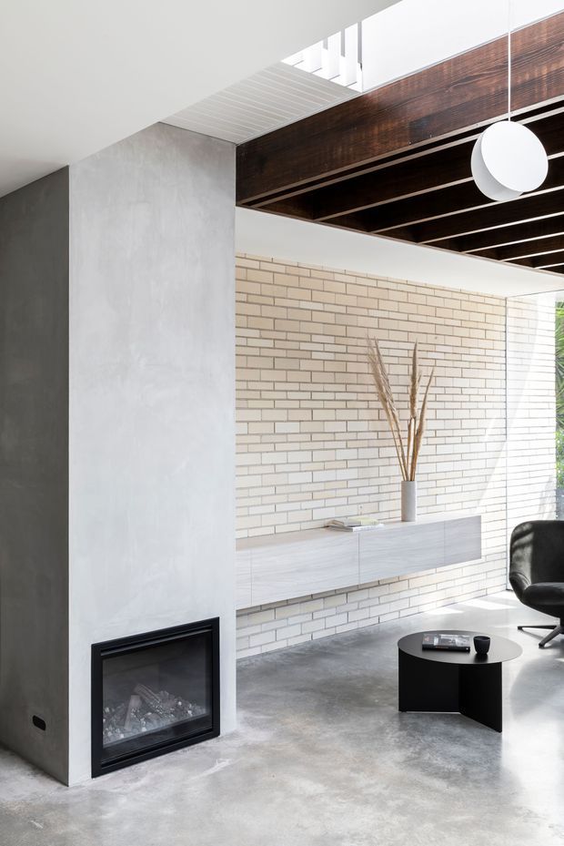 Bright contemporary living room with a minimalist built-in fireplace set into a smooth grey plastered chimney wall. A pale horizontal brick feature wall spans the room behind a long floating timber-toned console, while dark exposed ceiling beams contrast with white ceiling planes. The space includes a black lounge chair, low round black coffee table, sculptural white pendant light, and restrained natural decor.