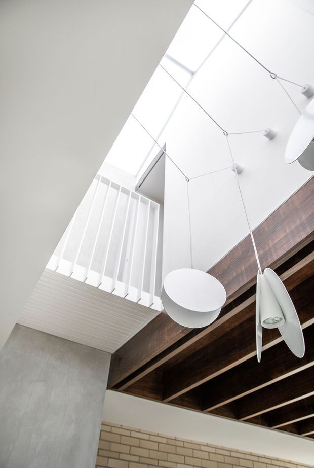 Upward-looking interior view of a bright double-height space with a large skylight or clerestory, white balustrade, exposed dark timber ceiling beams, suspended sculptural pendant lights, a smooth grey plaster wall, and a light brick wall below. The image emphasises the ceiling structure, natural light, and vertical volume.
