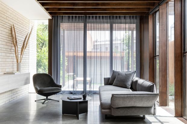 A refined contemporary living room with a grey sofa and sculptural armchair arranged around a low black coffee table. The room features polished concrete flooring, a white brick fireplace wall, extensive floor-to-ceiling glazing with sheer curtains, dark timber-framed openings, and an exposed timber beam ceiling. Bright natural light creates a calm, sophisticated atmosphere.