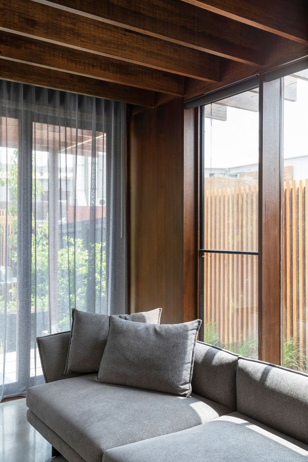 A close-up interior view of a calm sitting area featuring a large grey upholstered sectional sofa with cushions, set beside full-height glazed windows and sheer charcoal curtains. The room has rich dark timber wall panelling and exposed timber ceiling beams, with strong natural daylight and views to a timber privacy fence and planted outdoor area beyond.