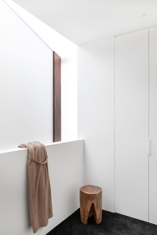 Minimalist white interior corner with full-height built-in cabinetry, a large frameless mirror or reflective panel, a low white half-height partition, and dark carpet flooring. A taupe towel hangs over the partition beside a small rustic timber stool, creating a restrained, monochromatic dressing or wardrobe area.